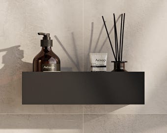 May include: A matte black rectangular wall-mounted shelf holding an amber glass bottle with a black pump dispenser labeled Aesop, a small white tube labeled Aesop, and a glass reed diffuser with black reeds against a light textured wall.