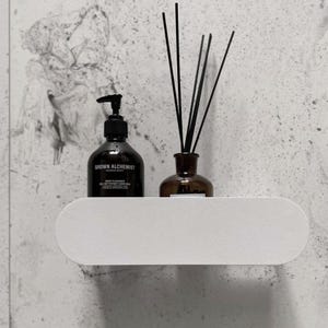 Könnte beinhalten: Ein weißes, ovales Wandregal mit einer schwarzen Pumpflasche mit der Aufschrift Grown Alchemist Body Cleanser und einer braunen Glasflasche mit schwarzen Diffusorstäbchen. Das Regal hat eine glatte, matte Oberfläche und eine längliche Form.