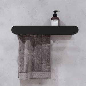 May include: A black metal shelf with rounded edges, mounted to a wall. A brown hand towel hangs from the shelf, and a bottle of hand soap sits on the shelf. The soap bottle is labeled "LA BRUKET HAND SOAP".