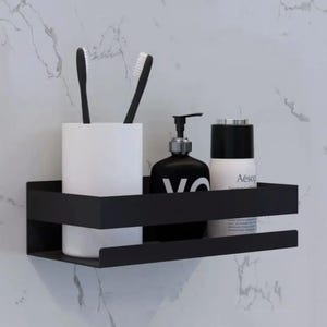 May include: Black metal bathroom shelf with two tiers. The shelf is holding a white ceramic toothbrush holder with two black toothbrushes, a black bottle with a pump dispenser, and a black and white bottle with the text "Aesop" on the label.