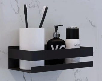 May include: A matte black rectangular metal shower caddy shelf holding a white cylindrical toothbrush holder with two black toothbrushes, a black pump bottle with white text, and a white cylindrical bottle with the text Aesop printed on it.