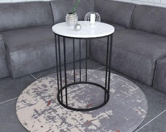 May include: A round side table with a white top and a black metal wire frame base. The table features a cylindrical structure with vertical black metal bars. On the tabletop sits a small silver sphere and a white candle inside a wire cage.