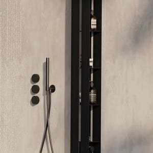 May include: A black, vertical shower storage unit with built-in shelves and a hanging loofah. The unit is mounted on a light grey tiled wall, next to a shower head and controls. Several brown bottles are visible on the shelves.