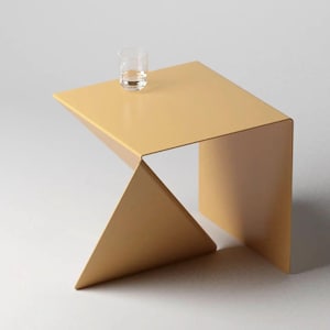 Puede incluir: Una mesa auxiliar de metal dorado con un diseño geométrico. La mesa tiene una tapa cuadrada y una base triangular.