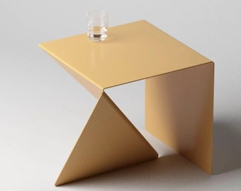 May include: A light yellow metal side table with a geometric, angular design featuring a flat square top and a triangular base support. A clear glass tumbler sits on the surface of the table, which is constructed from a single folded sheet.