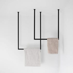Può includere: Un portasciugamani in metallo nero opaco montato a soffitto con tre aste di supporto verticali e due barre orizzontali. Due asciugamani rettangolari color beige chiaro sono drappeggiati sulle barre orizzontali.