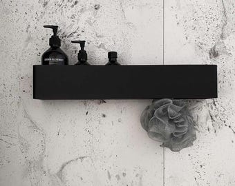 May include: A matte black rectangular wall-mounted shelf holding two dark pump bottles with labels and one small dark bottle. A grey mesh bath sponge hangs from the right side of the shelf. The shelf has a clean, solid, and minimalist design.