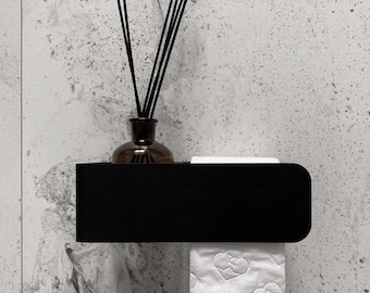May include: A matte black rectangular wall-mounted shelf featuring a flat top surface and a lower integrated roll holder. A dark glass diffuser bottle with black reed sticks sits on the shelf, and a roll of white toilet paper hangs below.