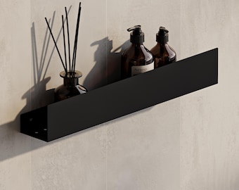 Può includere: Una mensola da parete nero opaco che sostiene un diffusore a bastoncini in vetro con bastoncini neri e due flaconi a pompa in vetro ambrato con etichetta Aesop. La mensola presenta un design rettangolare pulito con un pannello frontale.
