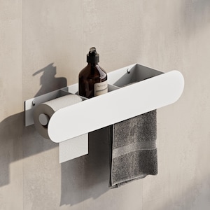Può includere: Un organizer da bagno in metallo bianco montato su una parete piastrellata di colore chiaro. Contiene un rotolo di carta igienica, una bottiglia marrone scuro e un asciugamano grigio. L'organizer ha un design curvo e un ripiano.