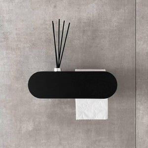 Puede incluir: Un portarrollos de papel higiénico negro, de forma ovalada, montado en la pared con una balda incorporada. El soporte tiene un rollo de papel higiénico blanco y un pequeño difusor con varillas negras. El diseño es moderno y minimalista.