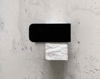 May include: A matte black rectangular wall-mounted toilet paper holder featuring a solid horizontal bar design. A single sheet of white toilet paper with a subtle embossed heart pattern hangs down from the center of the black metal fixture.