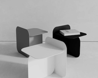May include: Three side tables in white, gray, and black, each featuring a flat square top and a curved vertical support panel. The black table has a rectangular book resting on its surface. The tables are arranged in a cluster on a flat surface.