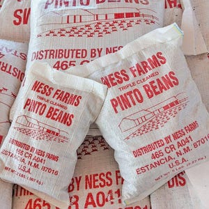 May include: Several white burlap sacks of pinto beans are stacked together. The bags have red text that reads "NESS FARMS PINTO BEANS" and "DISTRIBUTED BY NESS FARMS". The bags are labeled with the weight in pounds and location.
