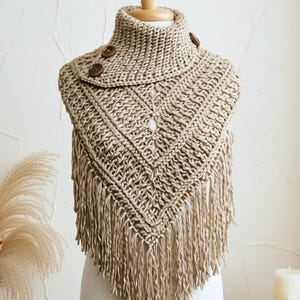 May include: A beige crochet cowl scarf featuring a triangular front drape with long fringe detailing. The garment has a high neck collar secured with two dark brown circular buttons on the side, showcasing a textured knit pattern throughout.