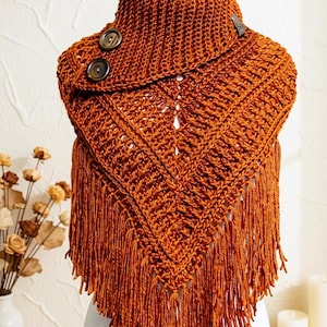 May include: A burnt orange crochet cowl scarf featuring a triangular front drape with long fringe detailing. The garment is secured at the neck with two large, dark brown circular buttons, showcasing a ribbed knit texture throughout the piece.