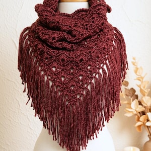May include: A deep burgundy triangular crochet shawl featuring an intricate openwork stitch pattern and long, thick fringe along the bottom edges. The garment is draped around a mannequin, showcasing the textured yarn construction and the pointed shape.