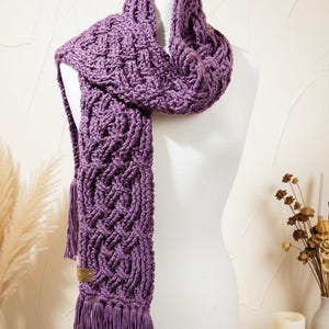 May include: A purple crocheted scarf featuring an intricate braided cable pattern throughout the length of the fabric. The scarf includes a small rectangular brown leather tag near the bottom edge and is finished with long fringe tassels.