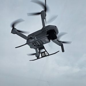 Puede incluir: Un dron gris con cuatro hélices está volando en el aire contra un cielo nublado. El dron tiene una cámara en la parte inferior y un tren de aterrizaje.