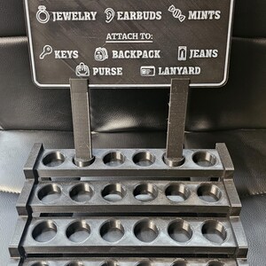 May include: A black 3D-printed tumbler keychain display. The sign reads "Tumbler Keychain" and lists items like cash, chapstick, and jewellery. It also lists places to attach the keychain, such as keys, backpack, and purse.