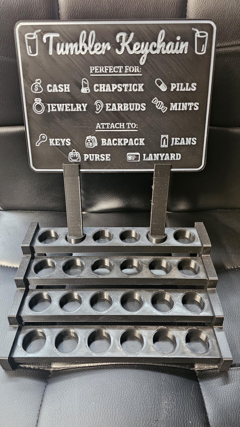 May include: A black 3D-printed tumbler keychain display. The sign reads "Tumbler Keychain" and lists items like cash, chapstick, and jewellery. It also lists places to attach the keychain, such as keys, backpack, and purse.