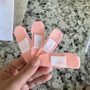 May include: Four pink felt bandages with white adhesive pads. The bandages are arranged in a fan shape, with the adhesive pads facing upwards. The background is a speckled gray surface.