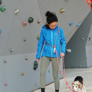 以下が含まれることがあります： 青いジャケットと緑色のレギンスを着た女性が、赤いリードでビーグル犬を連れて、クライミングウォールの前で散歩しています。