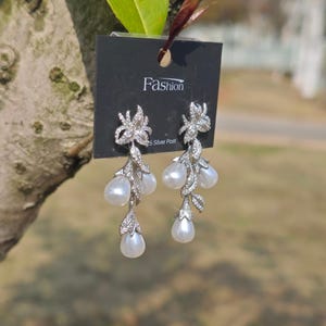 May include: Silver dangle earrings with a floral design and pearl drops. The earrings are on a black card with the word "Fashion" and "925 Silver Post" printed on it.