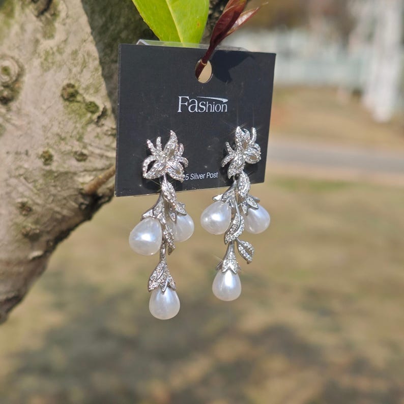 May include: Silver dangle earrings with a floral design and pearl drops. The earrings are on a black card with the word "Fashion" and "925 Silver Post" printed on it.
