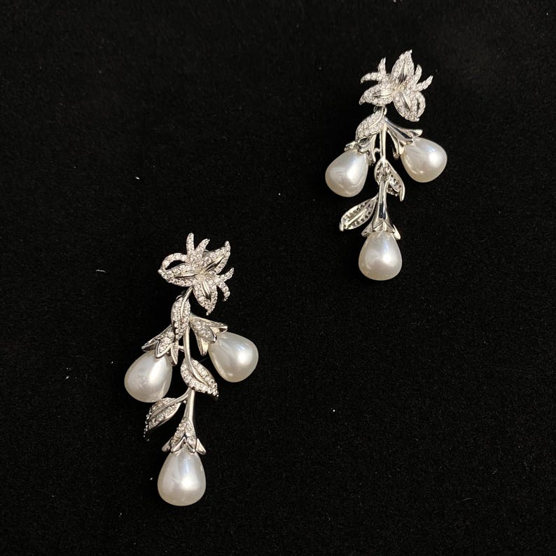 May include: A pair of silver earrings with a floral design and pearl drops. The earrings feature a delicate flower with sparkling stones and three pear-shaped pearls hanging from the stem.