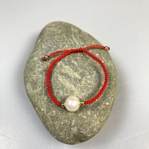 Op de afbeelding: Rode touw armband met een grote, witte parel kraal en kleine goudkleurige kralen. De armband is geknoopt en ligt op een grijze steen. Een eenvoudig, elegant sieraad.