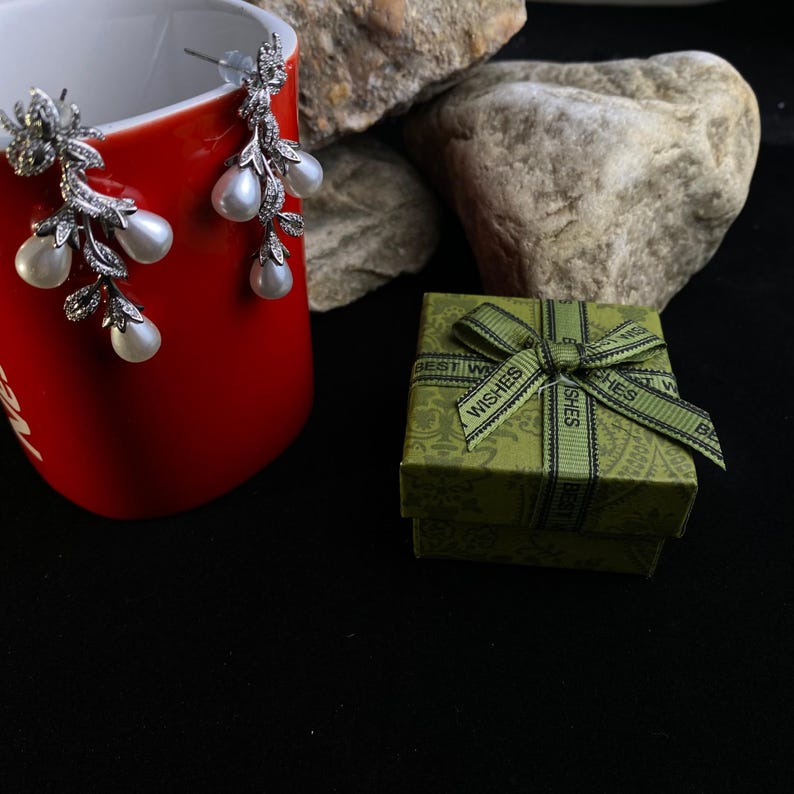 May include: A pair of silver dangle earrings with pearl accents, displayed on a red mug. The earrings are packaged in a green gift box with a black ribbon and the words "Best Wishes" printed on the box.