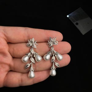 May include: Silver dangle earrings with pearlescent drops and a floral design. The earrings feature a cluster of three pearlescent drops, each with a delicate floral design.