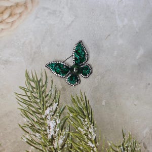 May include: A green jeweled butterfly brooch with a black pearl in the center.