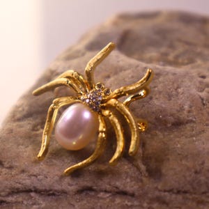 May include: A gold-toned spider brooch featuring a large, pale pink pearl. The spider's body is embellished with tiny, glittering gemstones. The brooch is displayed on a textured, grey stone surface.