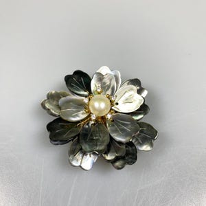 以下が含まれることがあります： グレーとシルバーの色合いの、虹色の花びらを持つ花のブローチ。中央には、小さな透明なクリスタルに囲まれた、大きくて丸いクリーミーな真珠があります。ブローチは金色の金属セッティングです。