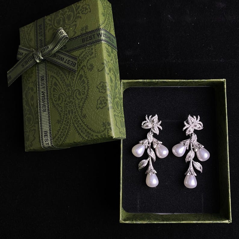 May include: A pair of silver dangle earrings with pearl drops and a floral design. The earrings are in a green gift box with a ribbon that says "Best Wishes".