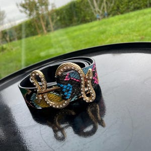 Könnte beinhalten: Ein schwarzer Gürtel mit einem bunten Schlangenmuster und einer goldenen Schlangenschnalle mit Strasssteinen.