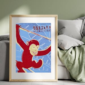 Puede incluir: Un póster vintage enmarcado con un mono rojo balanceándose en una línea amarilla y blanca sobre un fondo azul. El póster tiene texto en un idioma extranjero en la parte superior. El marco es de color madera clara.