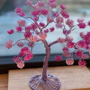Puede incluir: Una escultura de alambre rosa de un árbol con una base detallada e intrincada. El árbol tiene muchas ramas y hojas, todas hechas de alambre y pintadas de rosa.