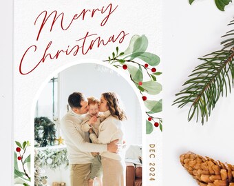 May include: A Christmas card with a watercolor design of green leaves and red berries. The card features a photo of a family of three, a man, a woman, and a child, with the text "Merry Christmas" and "The Reeds" in gold lettering. The date "DEC 2024" is printed in red.