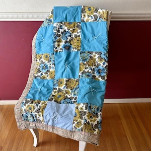 May include: A handmade quilt with a patchwork design. The quilt features squares of light blue fabric and floral patterns in blue, yellow, and white. The edges are trimmed with a patterned border. The quilt is draped over a white chair.