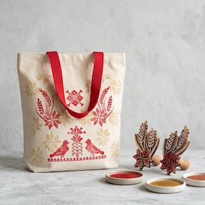 May include: A cream-coloured canvas tote bag with red fabric handles, featuring a red and gold cross-stitch pattern of birds, wheat stalks, and geometric shapes. Beside the bag are two wooden stamps with matching designs and three small paint dishes.