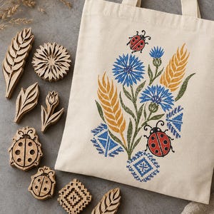 Op de afbeelding: Een beige canvas draagtas met een print van blauwe korenbloemen, gele tarwearen, rode lieveheersbeestjes en blauwe geometrische patronen, vergezeld door diverse uitgesneden houten stempels in de vorm van bladeren, bloemen en insecten.
