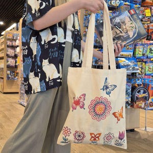 May include: A cream-coloured tote bag with colourful floral and butterfly designs. The bag has long, sturdy handles. The background shows a shop with toys and a Lego Harry Potter set.