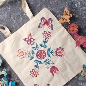 May include: A cream tote bag featuring a floral and butterfly pattern in pink, orange, and teal. Wooden butterfly and flower stamps and ink pads are arranged around the bag.