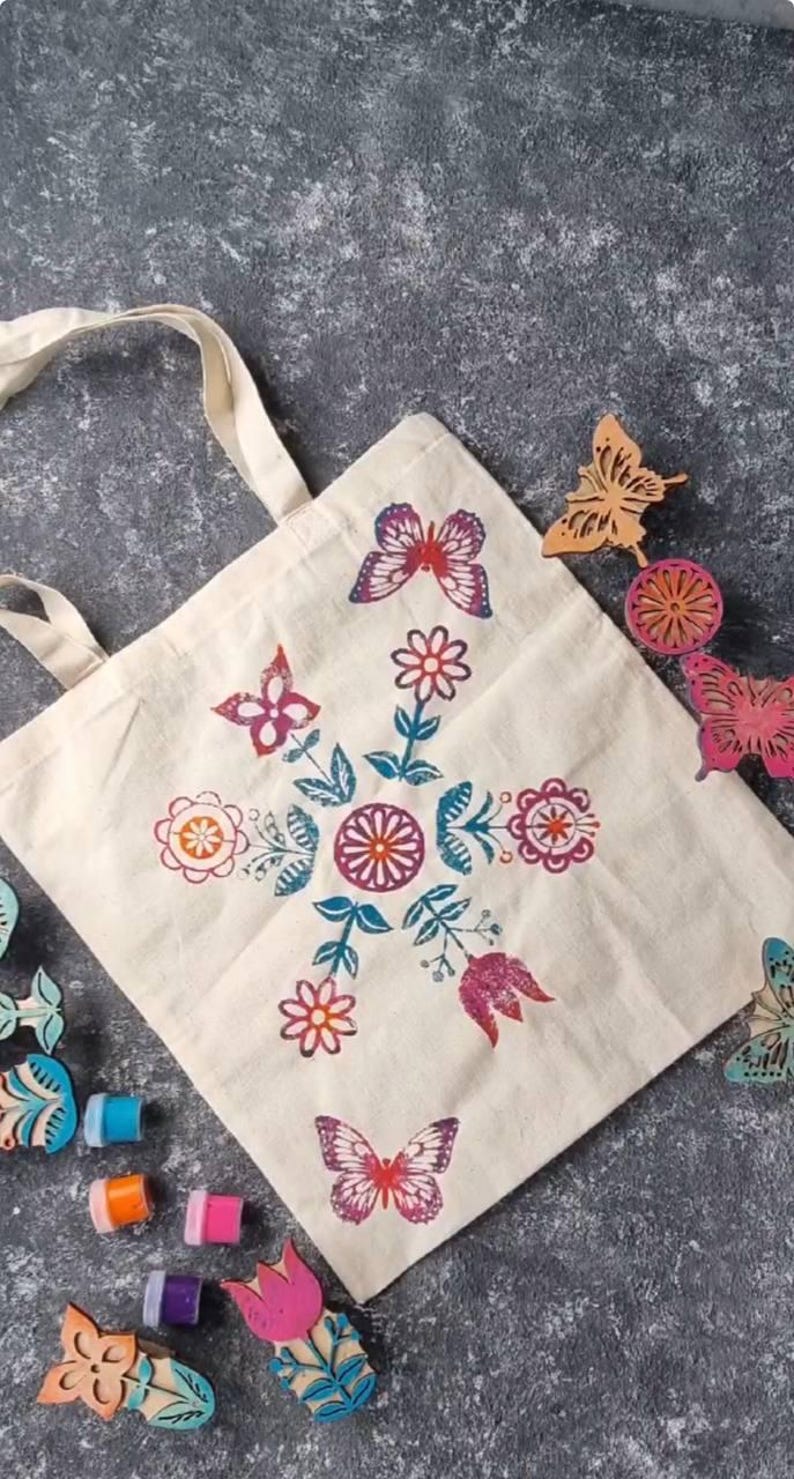 May include: A cream tote bag featuring a floral and butterfly pattern in pink, orange, and teal. Wooden butterfly and flower stamps and ink pads are arranged around the bag.