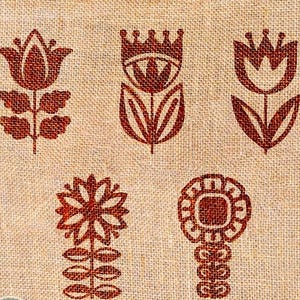 May include: Close-up of a beige fabric featuring a repeating pattern of reddish-brown floral designs. The flowers are stylized, with shapes including tulips and other geometric forms. The fabric has a textured appearance.