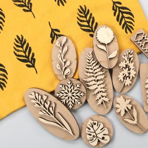 Może przedstawiać: Kolekcja drewnianych stempli z różnymi wzorami botanicznymi, w tym liśćmi, kwiatami i płatkiem śniegu. Stemple są owalne i prostokątne, z wyciętymi wzorami. Żółta tkanina z czarnymi wzorami liści w tle.