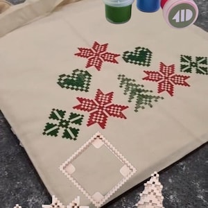 May include: A beige tote bag with red and green pixelated floral and heart designs. The image also shows small paint pots in yellow, blue, and green, along with wooden cutouts of snowflakes, hearts, and a Christmas tree, plus yellow sponge brushes.